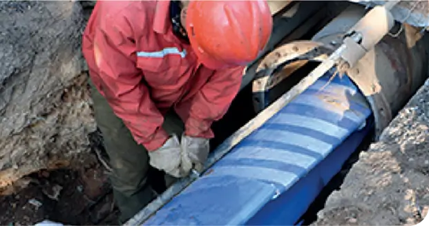 A technician in protective gear performing pipe rehabilitation on a large subterranean utility line within an excavated trench.
